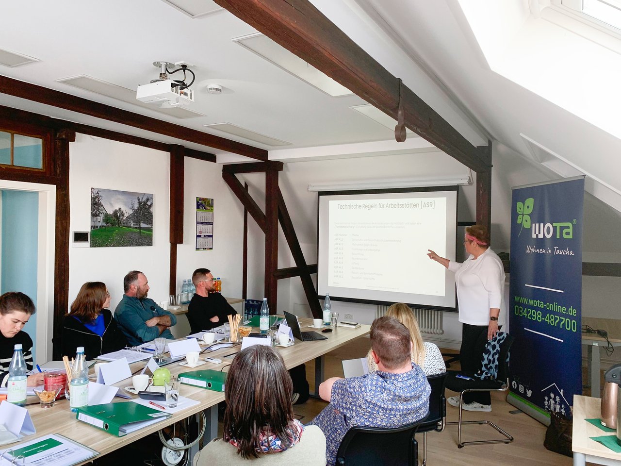 Schulung mit 7 Teilnehmenden, die vor einer Leinwand sitzen und der Schulungsleitung zuhören